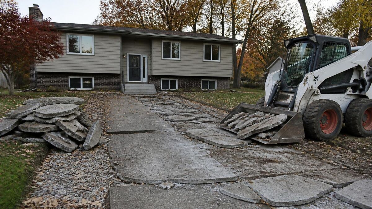 A skid steer breaking up a frost-heaved concrete driveway in front of a split-level house
