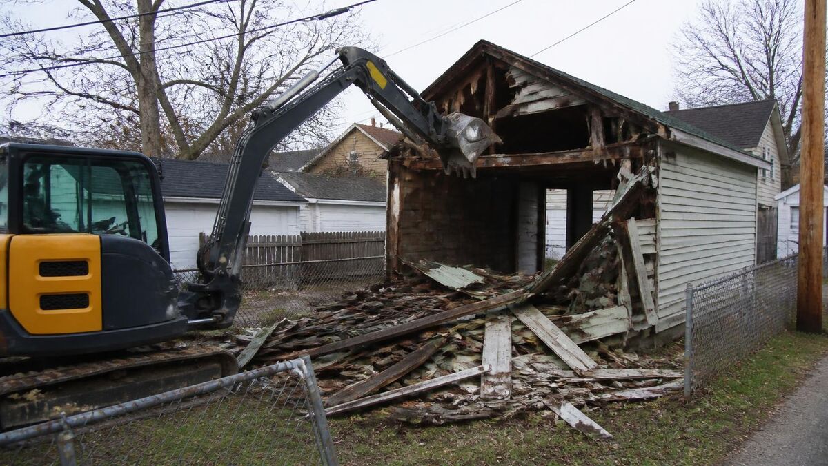 A mini excavator tearing down an old detached garage in a backyard next to a chain link fence