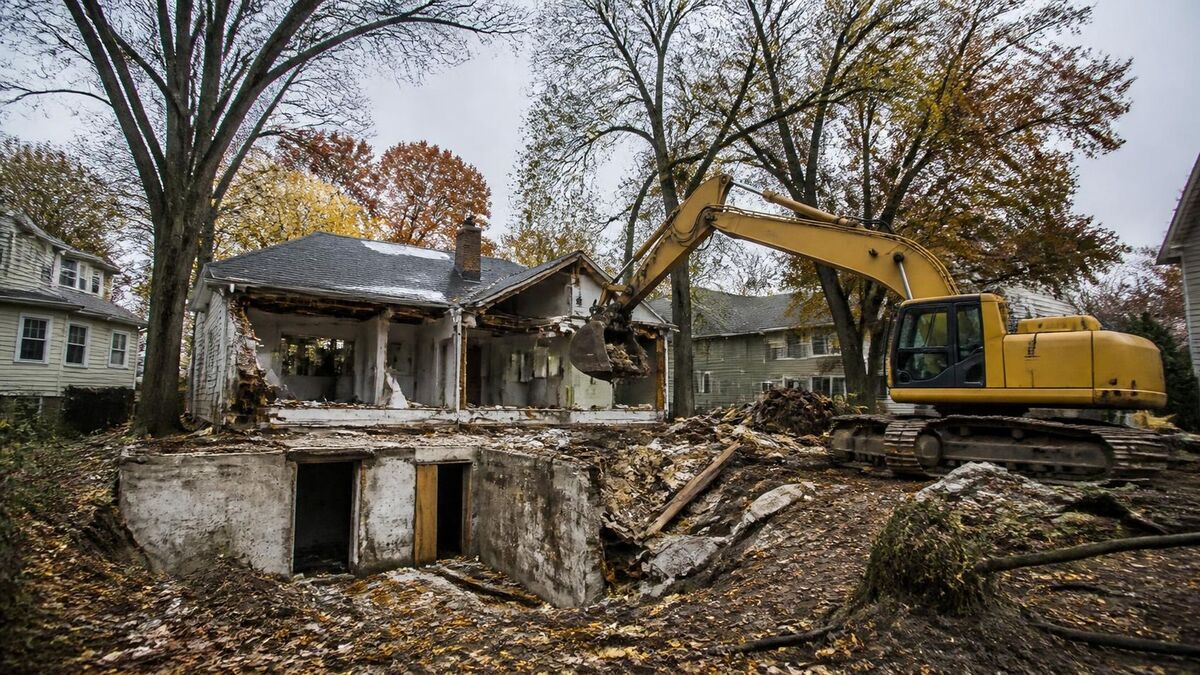 An excavator demolishing a rambler house down to its open basement among mature autumn trees