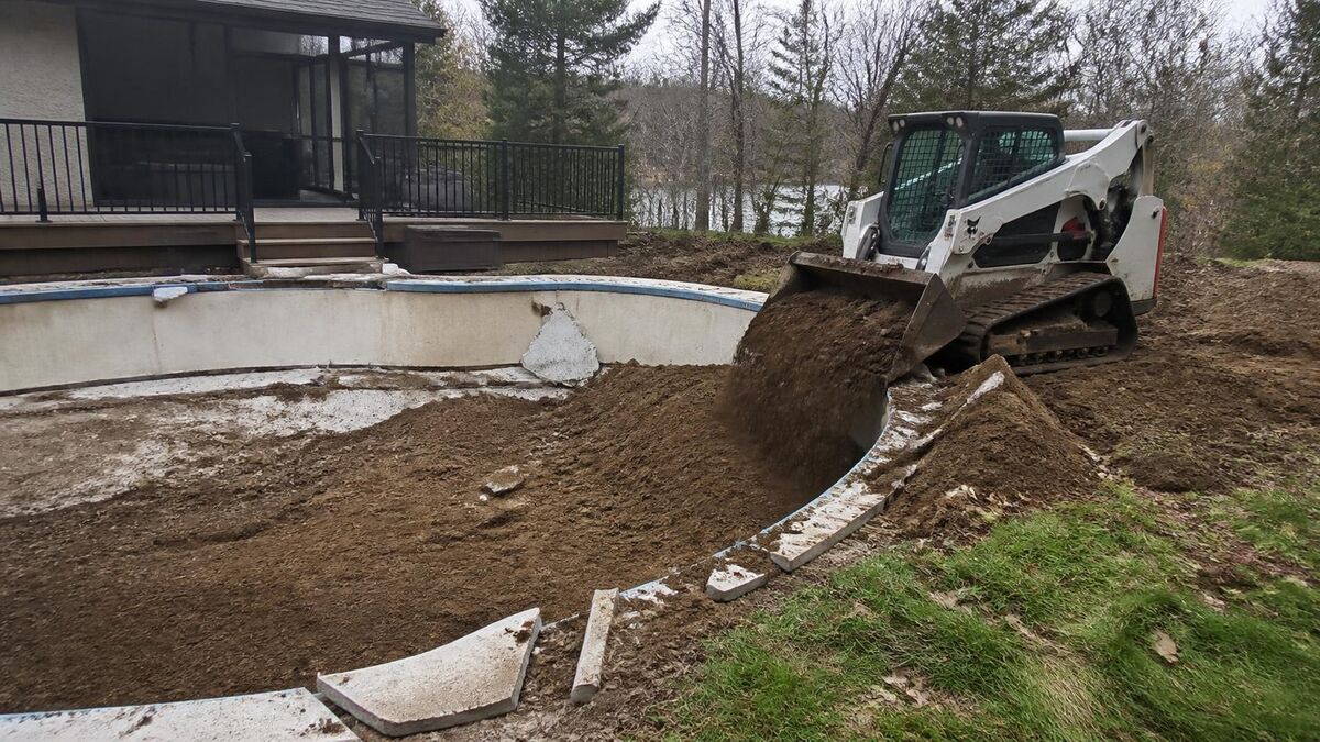 A compact track loader backfilling a broken-out in-ground pool in a wooded suburban backyard